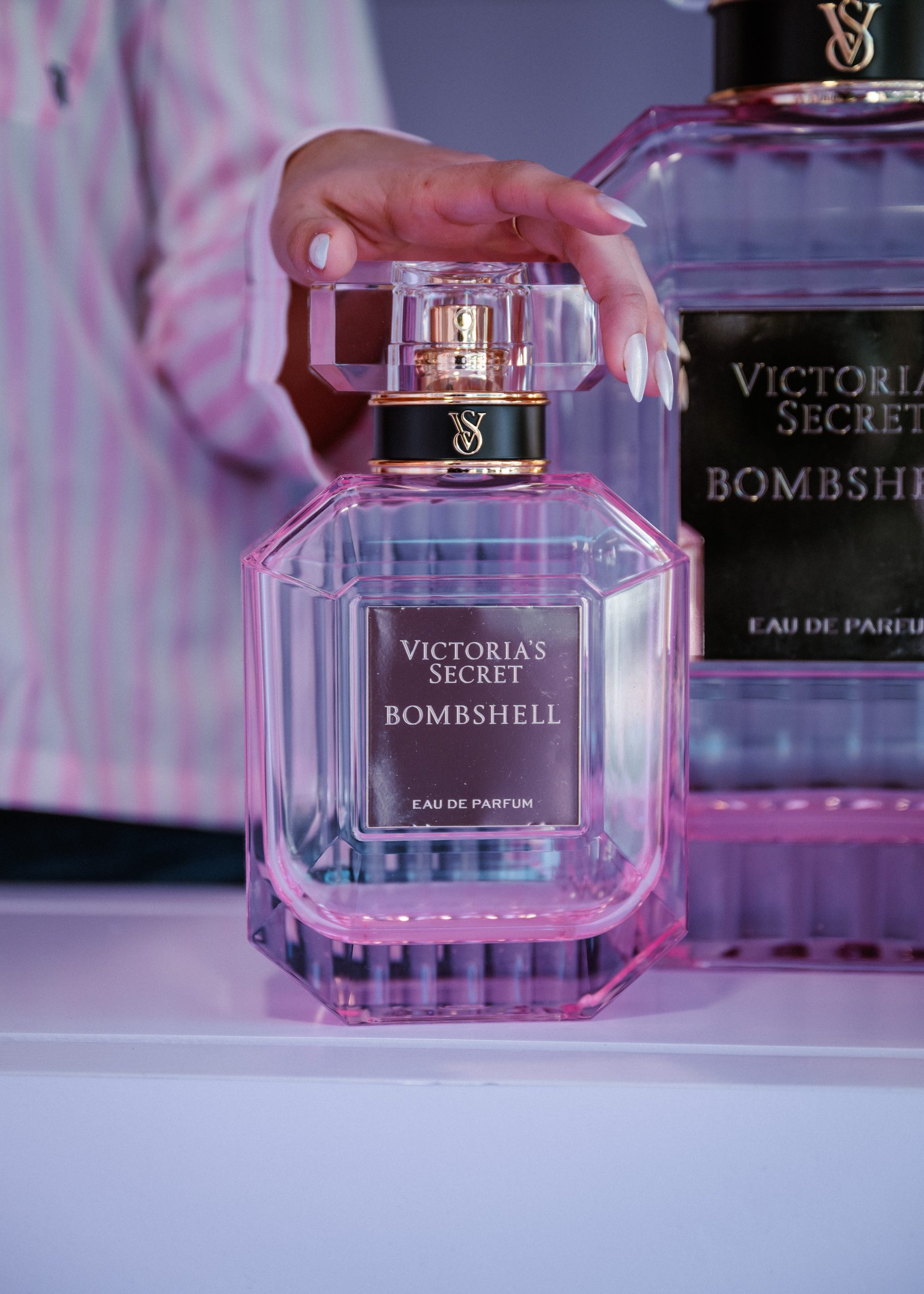 Hand with manicured nails holds a pink Victoria’s Secret Bombshell bottle; another bottle behind.