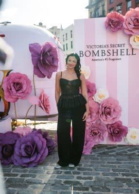 A woman in a black strapless top and black pants stands on a cobblestone street in front of a Victoria's Secret Bombshell display, surrounded by large pink and purple paper flowers.