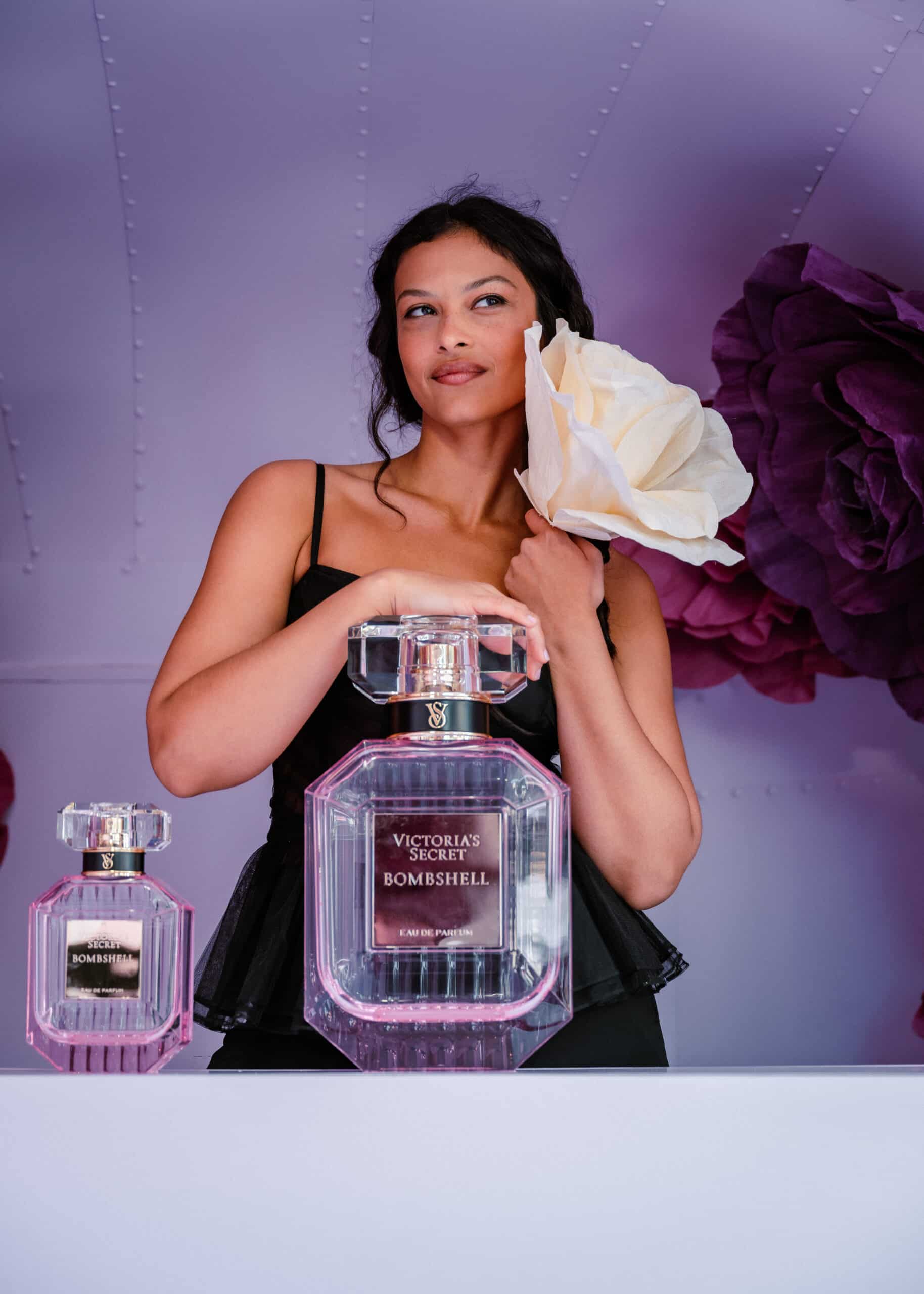 Woman in black dress behind counter with two large perfume bottles, holding big white paper flower.