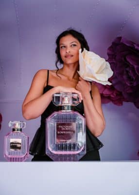 A woman in a black top holds a large white flower beside her face, standing next to two oversized Victoria’s Secret Bombshell perfume bottles against a purple background with large pink flowers.