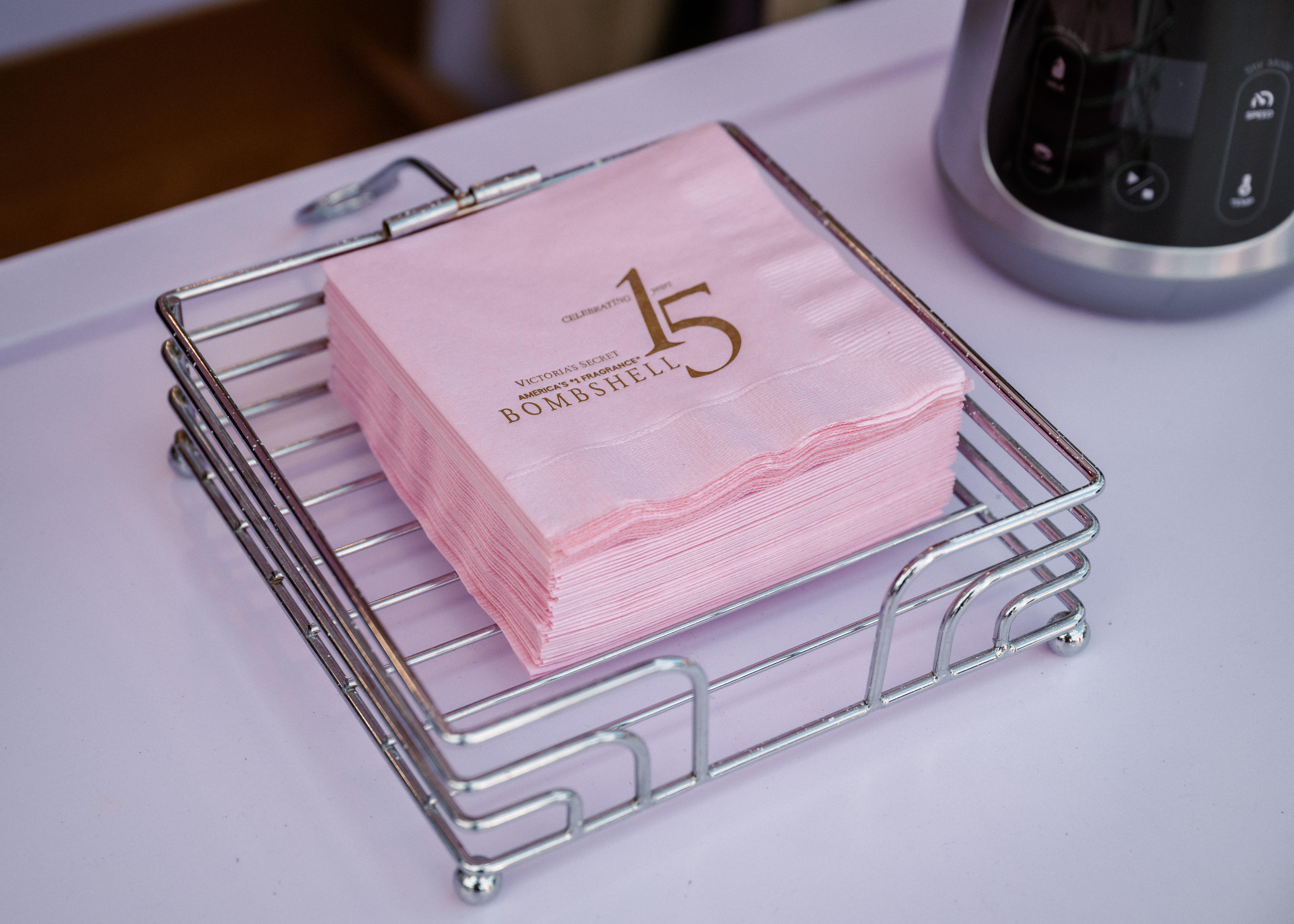 A stack of pink napkins in a silver holder on a white surface, with “15 Bombshell” printed in gold on the top napkin. Part of a coffee machine is visible on the right.