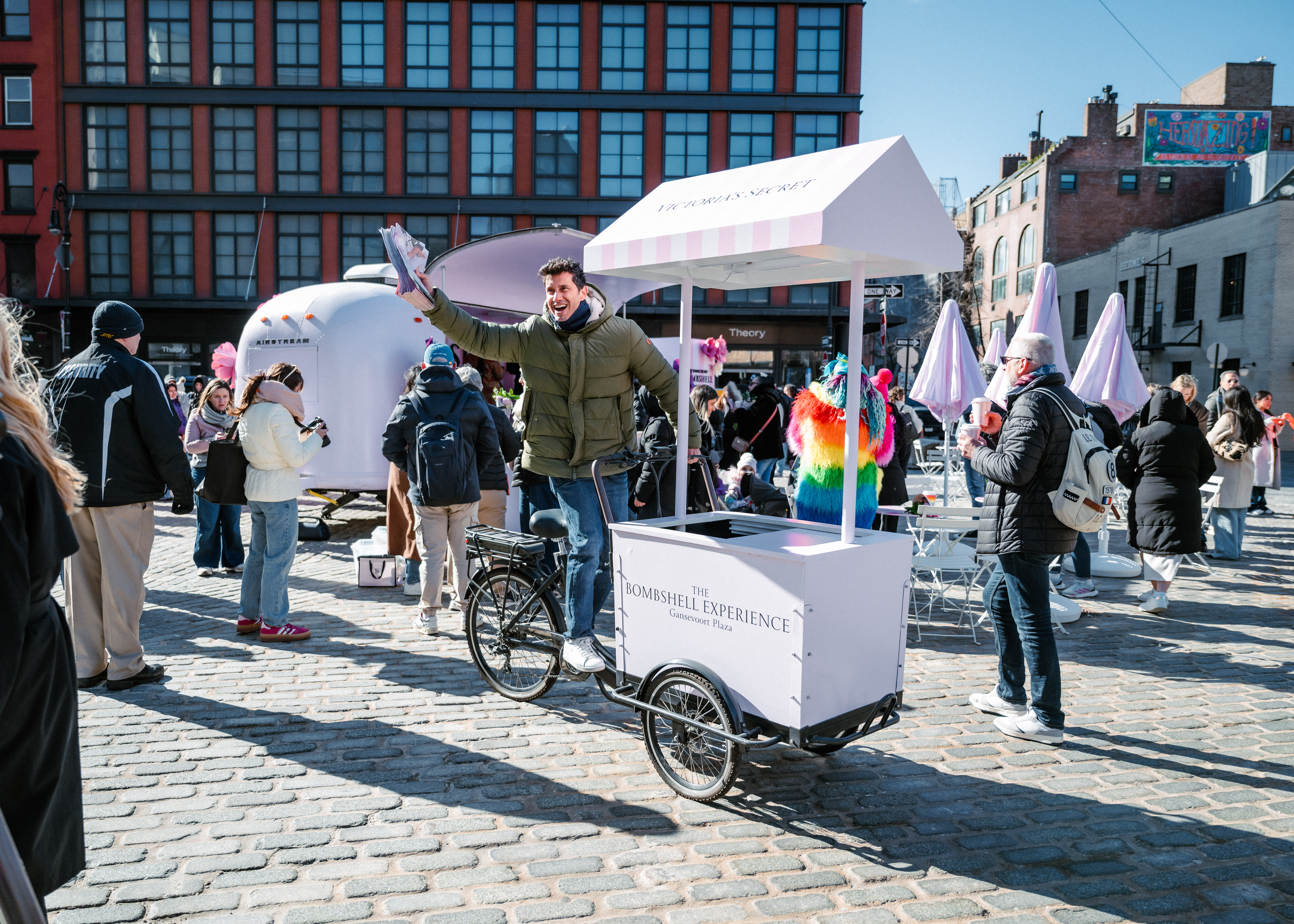 A man stands behind a white cart labeled "The Boardwalk Experience" on a cobblestone street, smiling and waving cotton candy. People gather around in a lively outdoor event with colorful decorations.