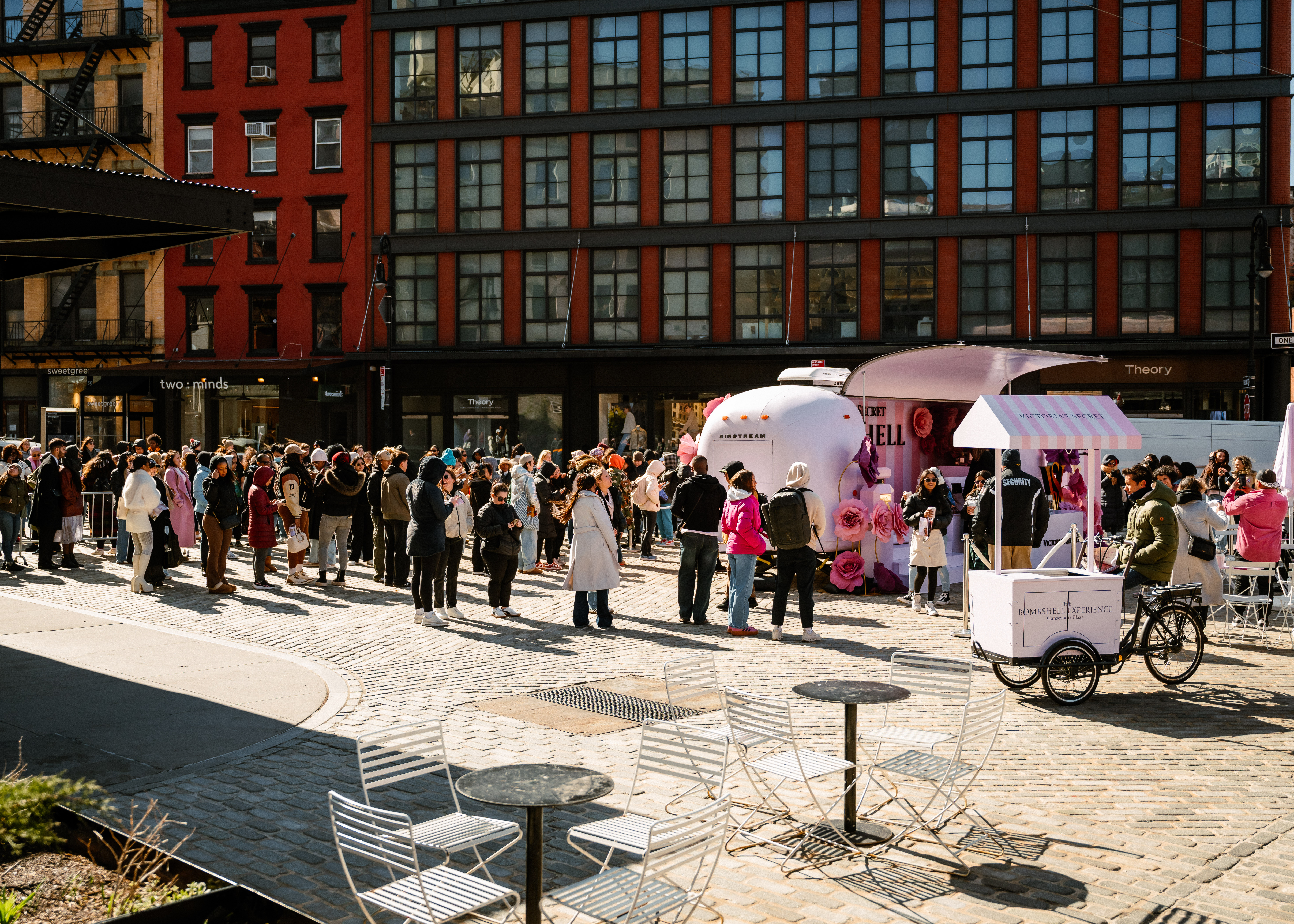 A large crowd waits in line at an outdoor event in a city plaza, with a bright pink booth and a small cart nearby. Modern buildings with many windows are in the background. Empty tables and chairs are in the foreground.