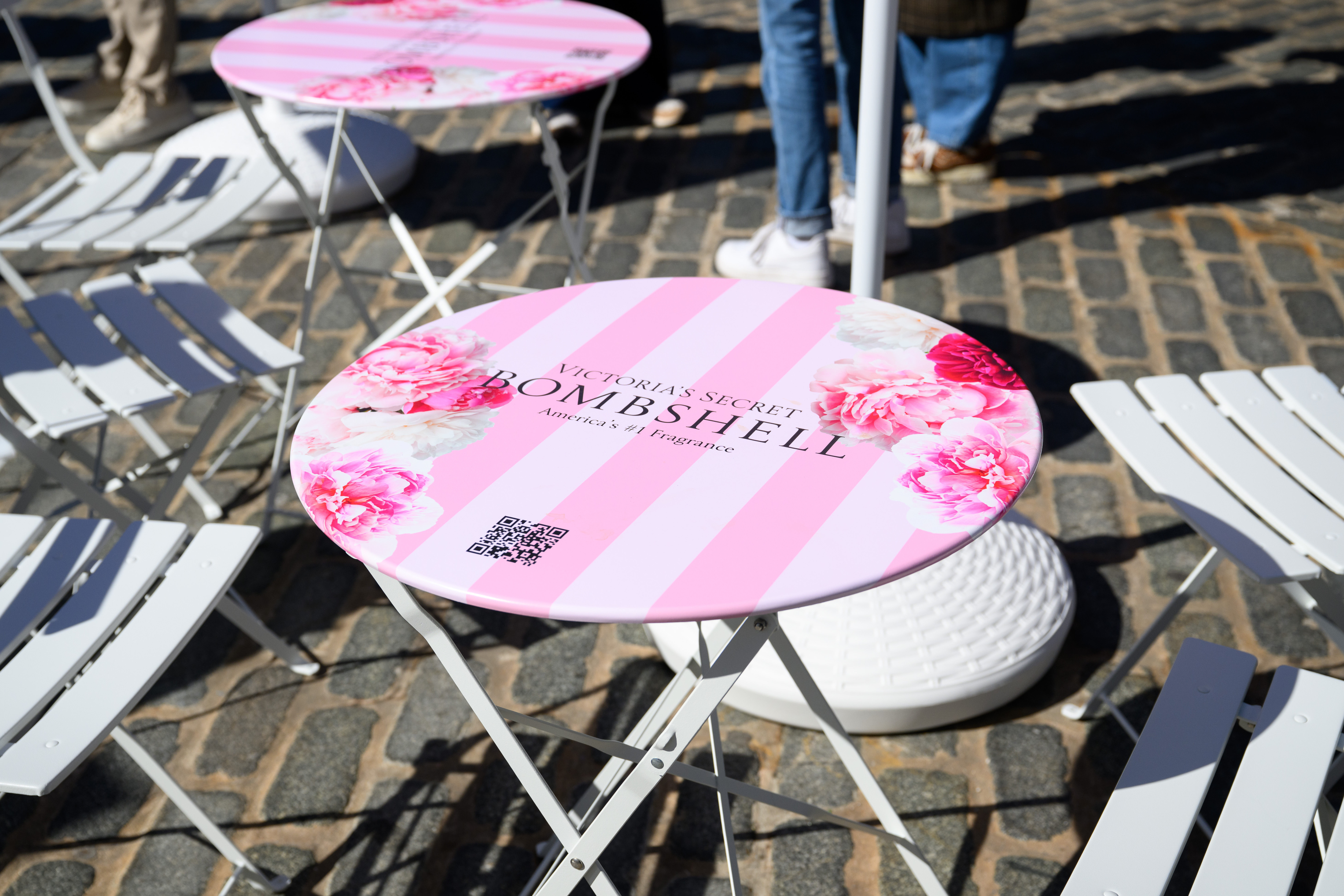 A round table with a pink striped Victoria’s Secret Bombshell design, floral accents, and a QR code, surrounded by white folding chairs on a cobblestone surface. People stand in the background.