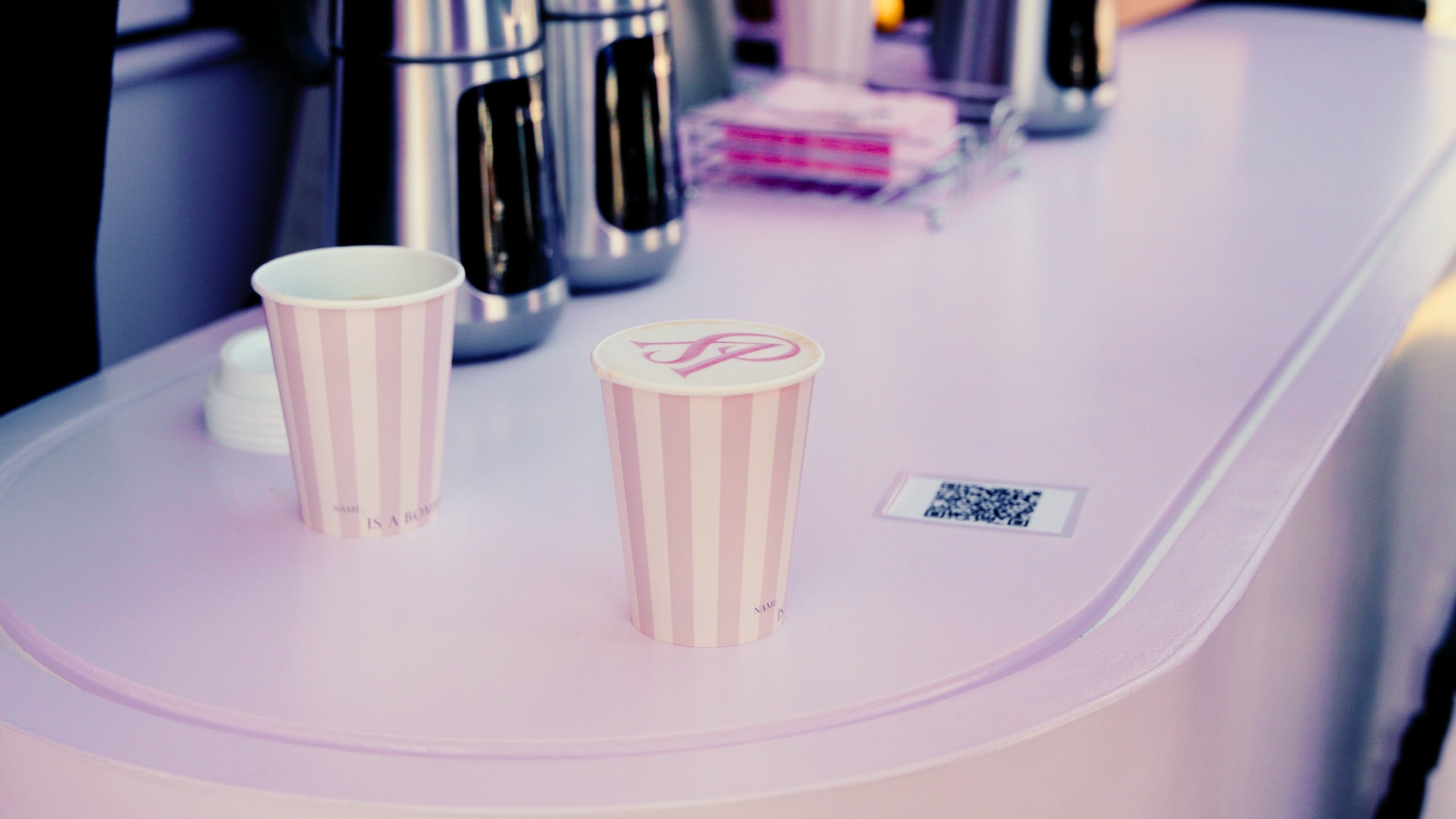 Two pink and white striped paper cups, one with a lid, sit on a light pink counter with a visible QR code. In the background, there are metal thermoses and stacked napkins.