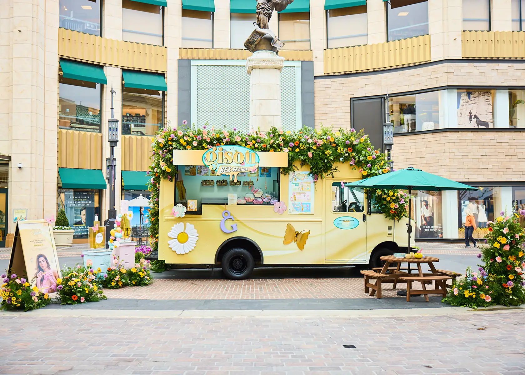 A pastel yellow flower-themed food truck, one of the vibrant experiential vehicles, is decorated with flowers and butterflies. Parked before a large building, it’s surrounded by blooms, a bench with an umbrella, and people strolling nearby.