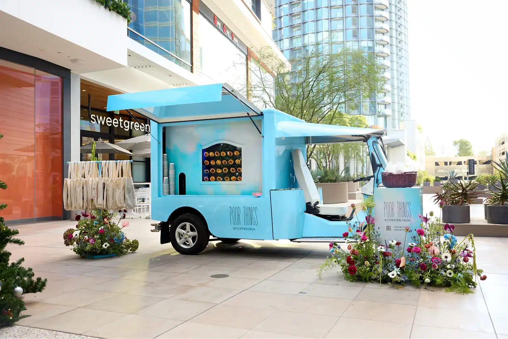 A light blue mobile marketing vehicle with a serving window is decorated with fresh flowers and parked in an outdoor mall area near Sweetgreen, with tote bags hanging outside and modern buildings in the background.