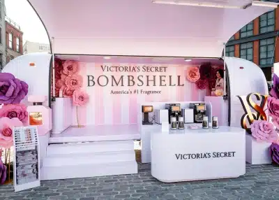 A Victoria’s Secret Bombshell fragrance pop-up display with pink striped walls, large paper flowers, and perfume bottles on white counters brings a touch of luxury to the city—perfect for experiential marketing trucks in an outdoor urban setting.