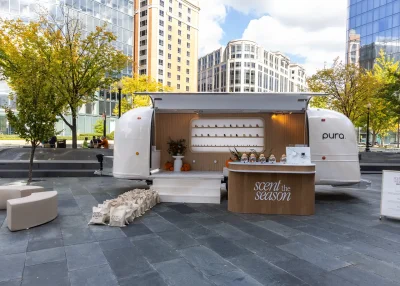 A white event marketing vehicle with a display of products and pumpkins is set up in an urban plaza. A wooden counter reads "scent the season," with gift bags arranged nearby and modern buildings amid autumn trees in the background.