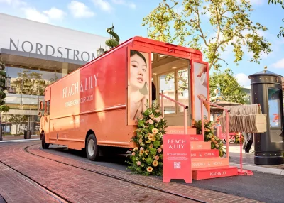 A bright peach-colored Peach & Lily skincare truck with floral decorations is parked outside a Nordstrom store. The stairs display product features, and a large image of a woman's face covers the side of the truck.