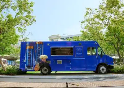 A bright blue food truck with skincare product ads on its side is parked outdoors among green trees. A small table with white flowers stands beside it, and buildings are visible in the background.