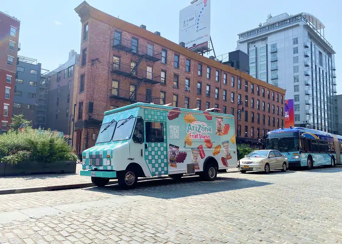 A colorful food truck with “AriZona Fruit Snacks” branding is parked on a cobblestone street next to a blue bus and a car, with brick and modern buildings in the background under a clear sky.