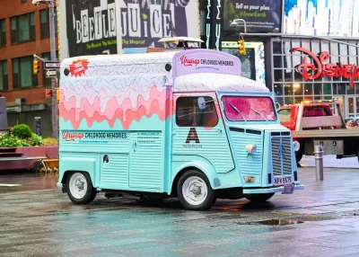 A pastel blue and pink ice cream van with “Childhood Memories” written on the side is parked on a wet city street, with bright billboards and Disney and Beetlejuice signs in the background.