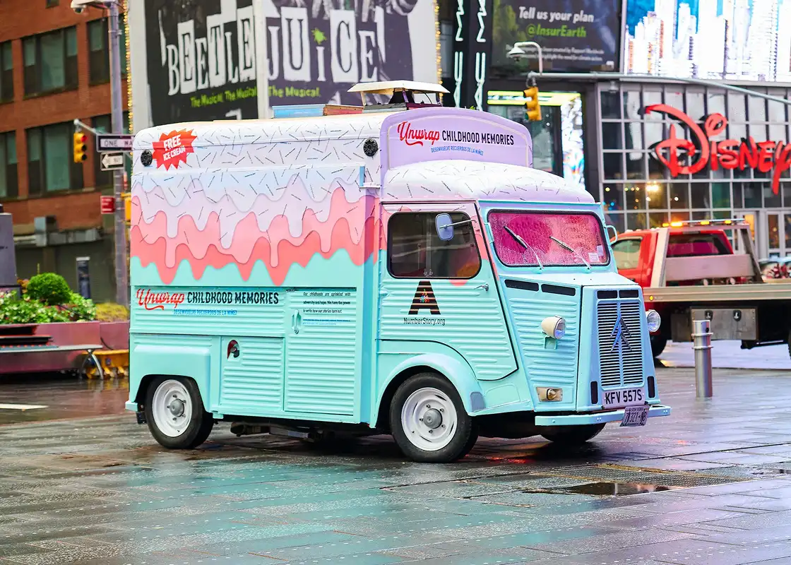 A pastel blue and pink ice cream van with “Childhood Memories” written on the side is parked on a wet city street, with bright billboards and Disney and Beetlejuice signs in the background.