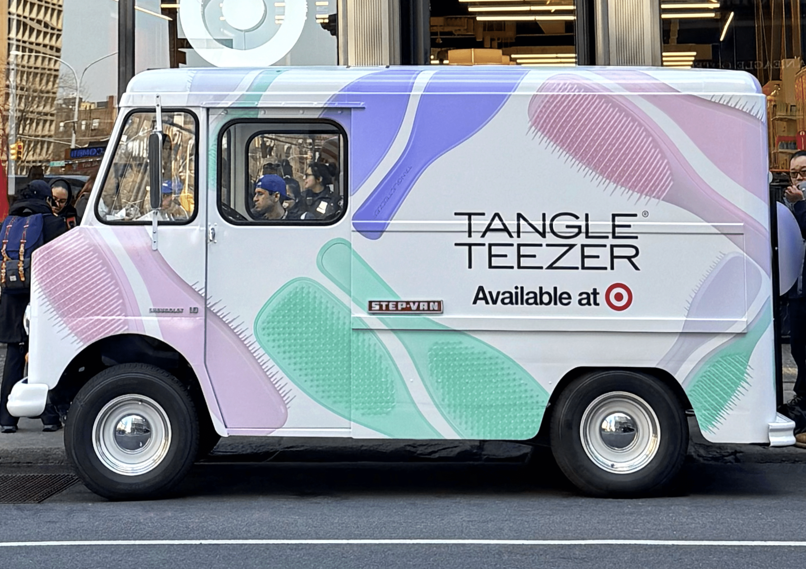 A delivery truck with Tangle Teezer branding is parked on a city street near shops and people.