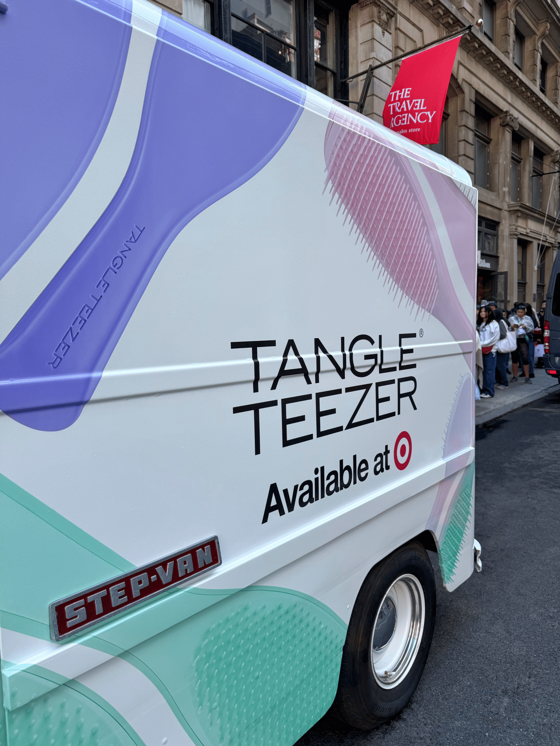 A van advertising Tangle Teezer at Target is parked by a building with a red Travel Agency flag.