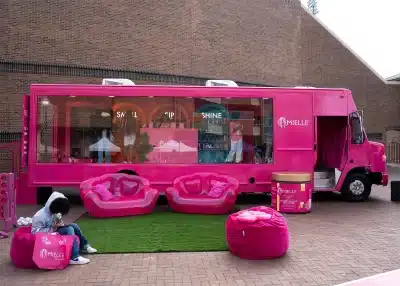 A bright pink Mielle-branded promotional vehicle with large windows is parked outside. In front, there are pink lounge chairs, bean bags, and a person sitting on a bean bag. Shelves with products are visible inside the truck.