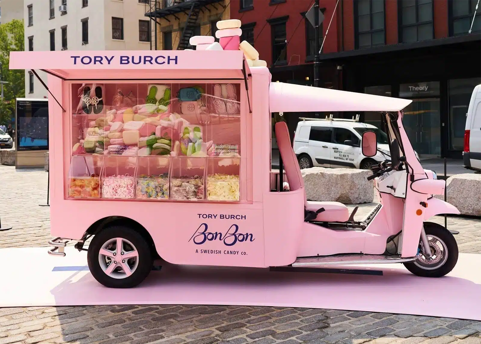 A pink three-wheeled cart labeled "Tory Burch BonBon" displays assorted colorful candies in clear bins. This eye-catching promotional vehicle features a candy-themed design and is parked outdoors on a cobblestone street.