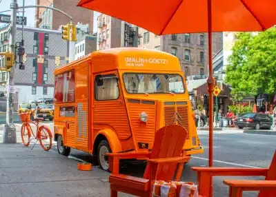 A bright orange vintage Citroën van with "MALIN+GOETZ new york." branding is parked on a city street. Orange chairs, a bike, and an umbrella are nearby, creating a vibrant, coordinated outdoor scene.