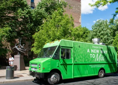 A bright green branded food truck with "A New Way To Mix" written on its side is parked on a city street near trees and a statue. A man stands next to the statue under the sunny sky.