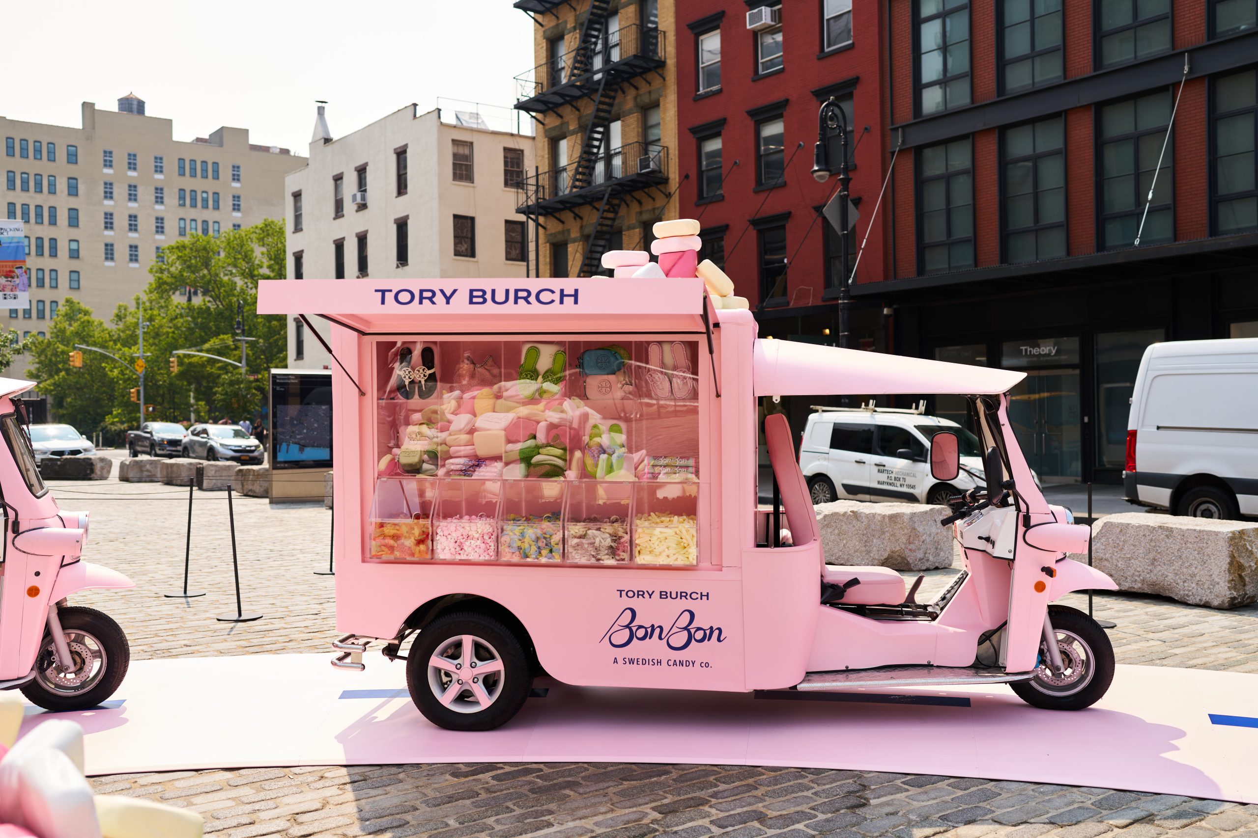 Pink Tory Burch BonBon food truck with candies parked on cobblestone street, buildings behind.