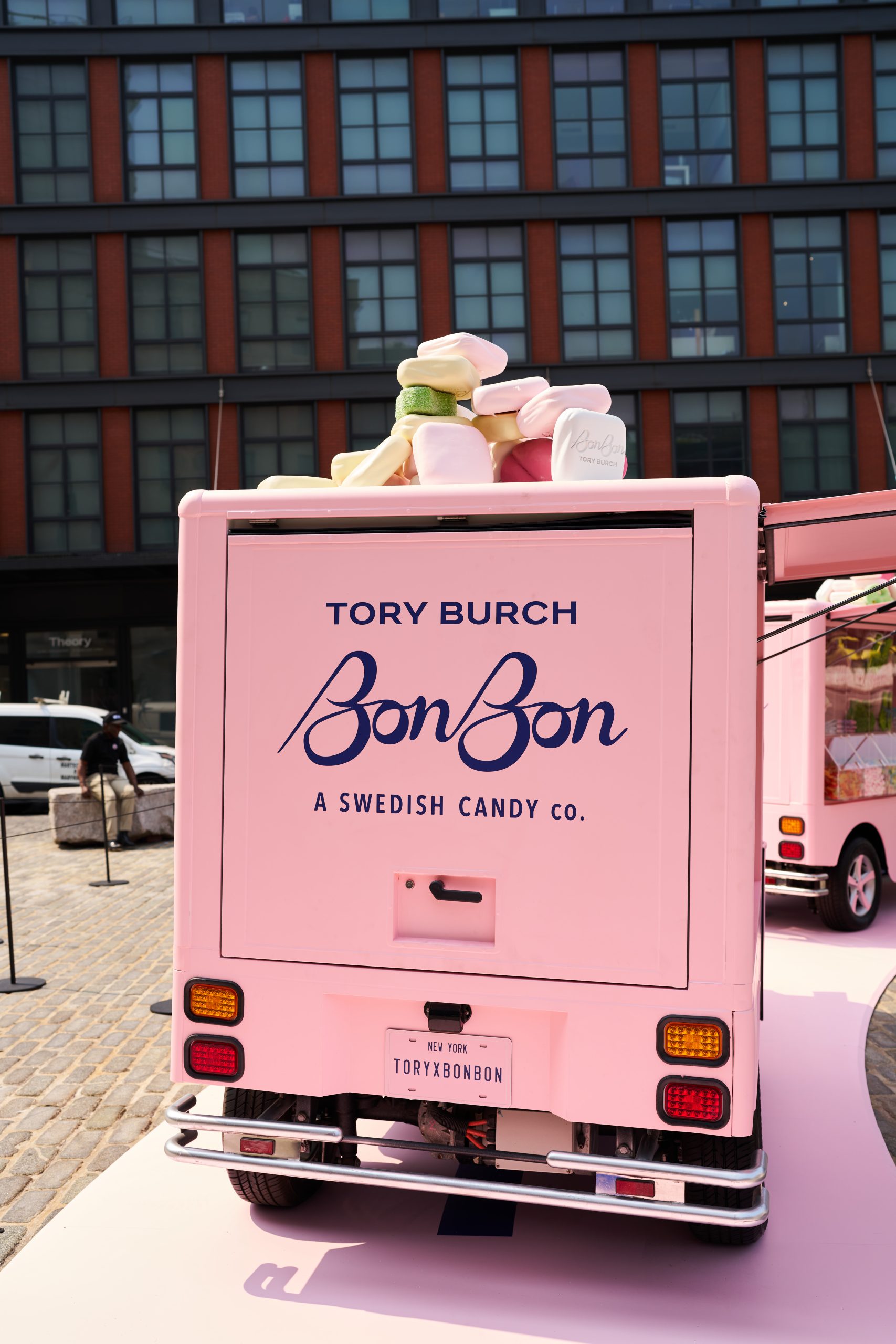 Pink food truck labeled Tory Burch BonBon with large candy props parked on cobblestone by building.
