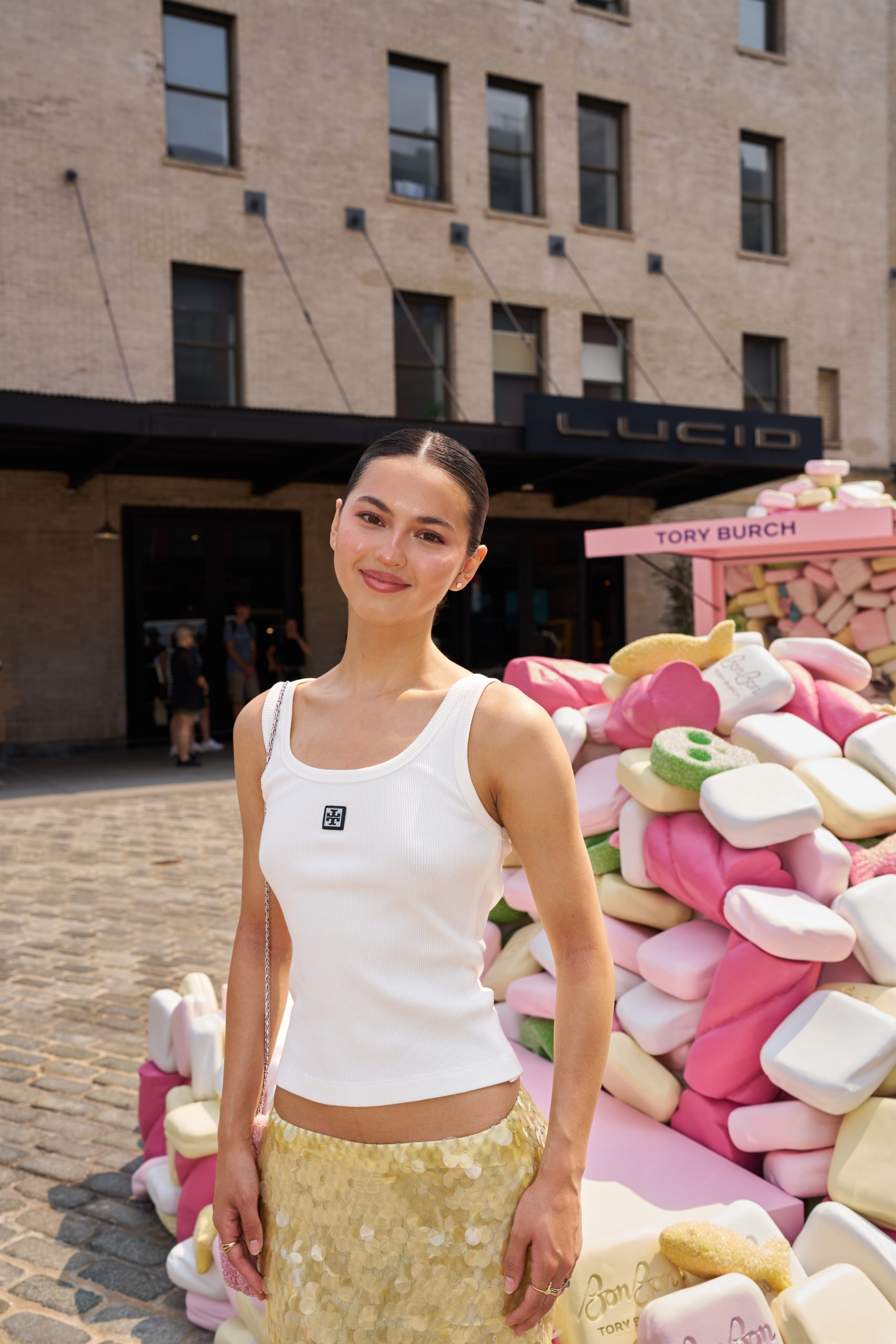 Woman in white tank top and gold sequin skirt stands by stacked pastel soaps, LUCID sign behind.