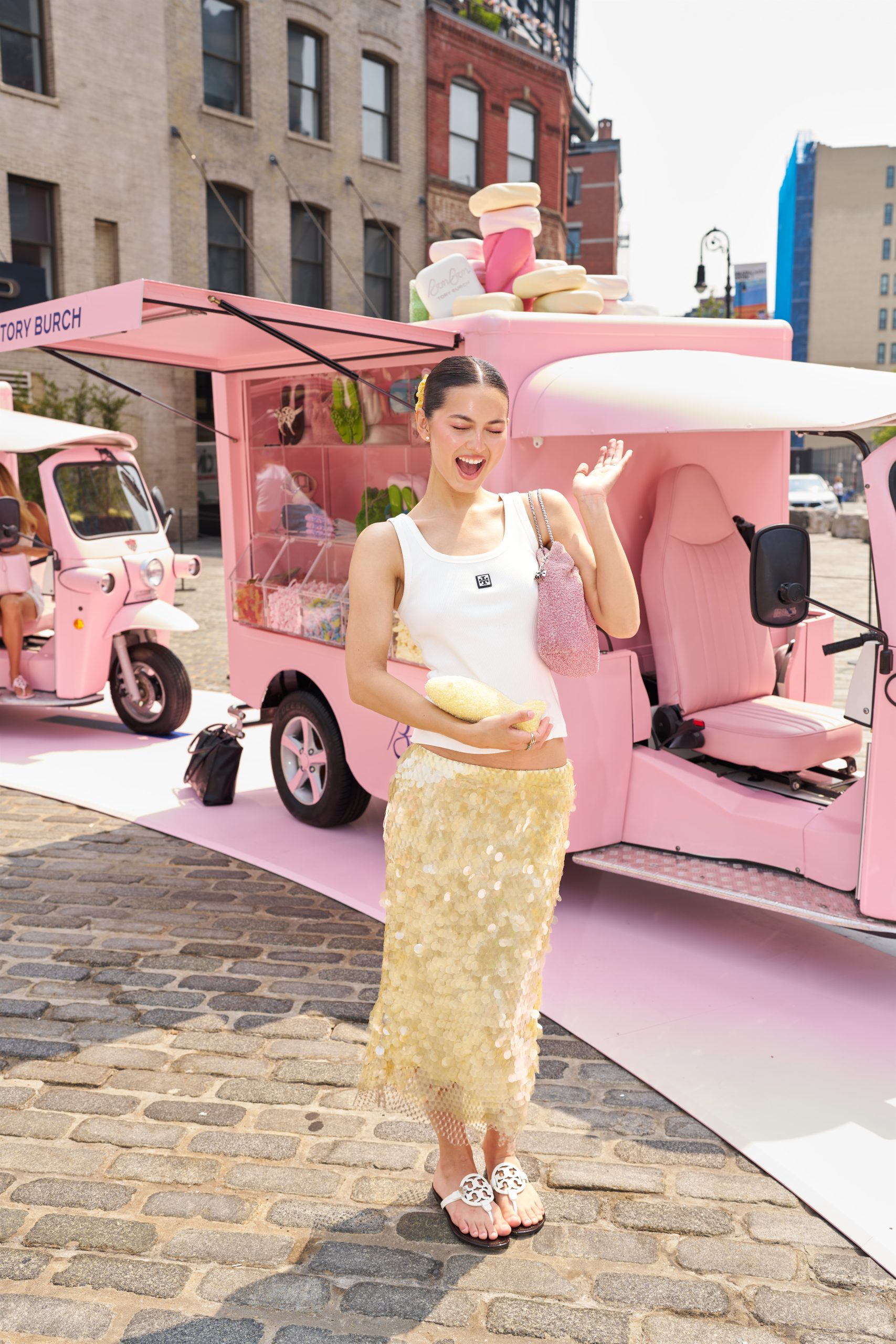 Woman in white tank top and yellow skirt waves by pink mobile shop selling hats and accessories.