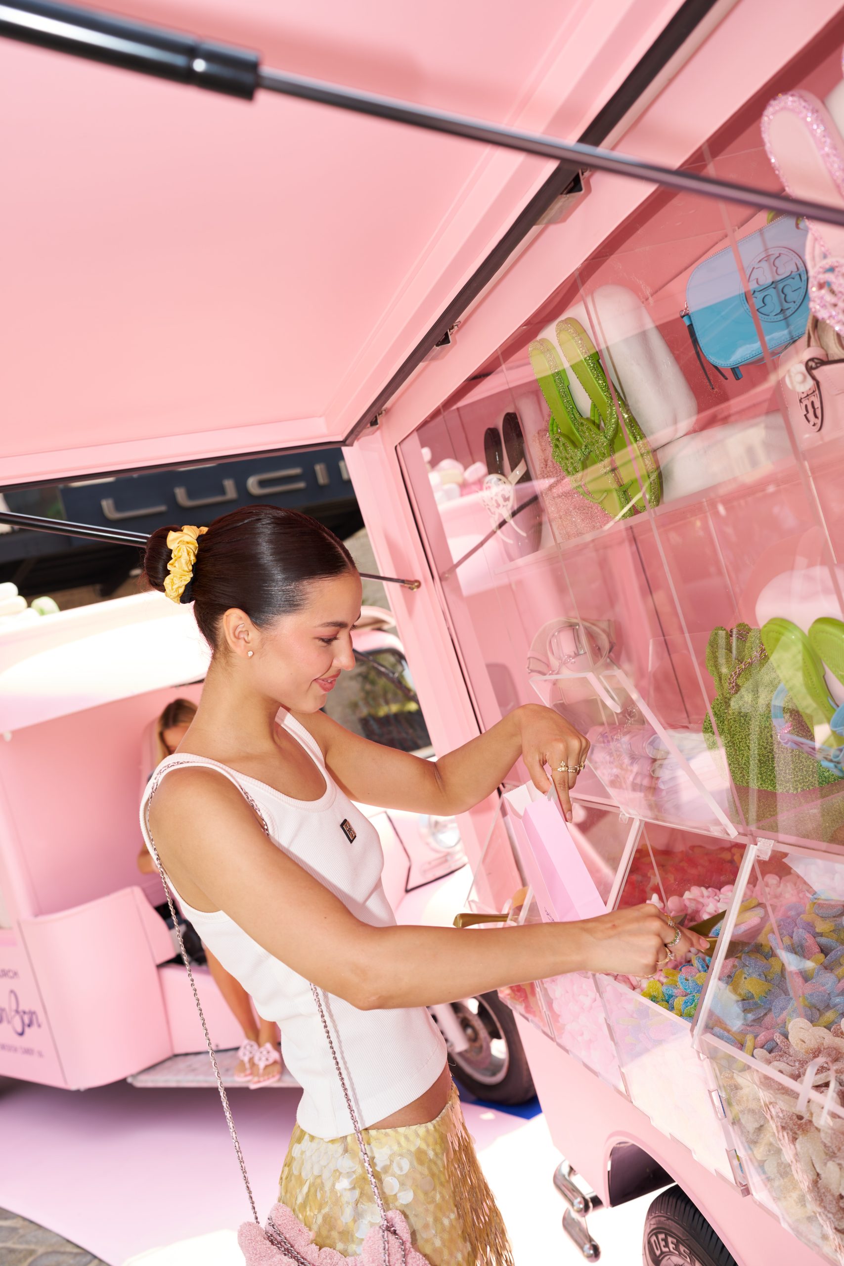 Woman with dark hair in a bun picks candy from clear bins on a pink cart with sweets and green flip-flops.