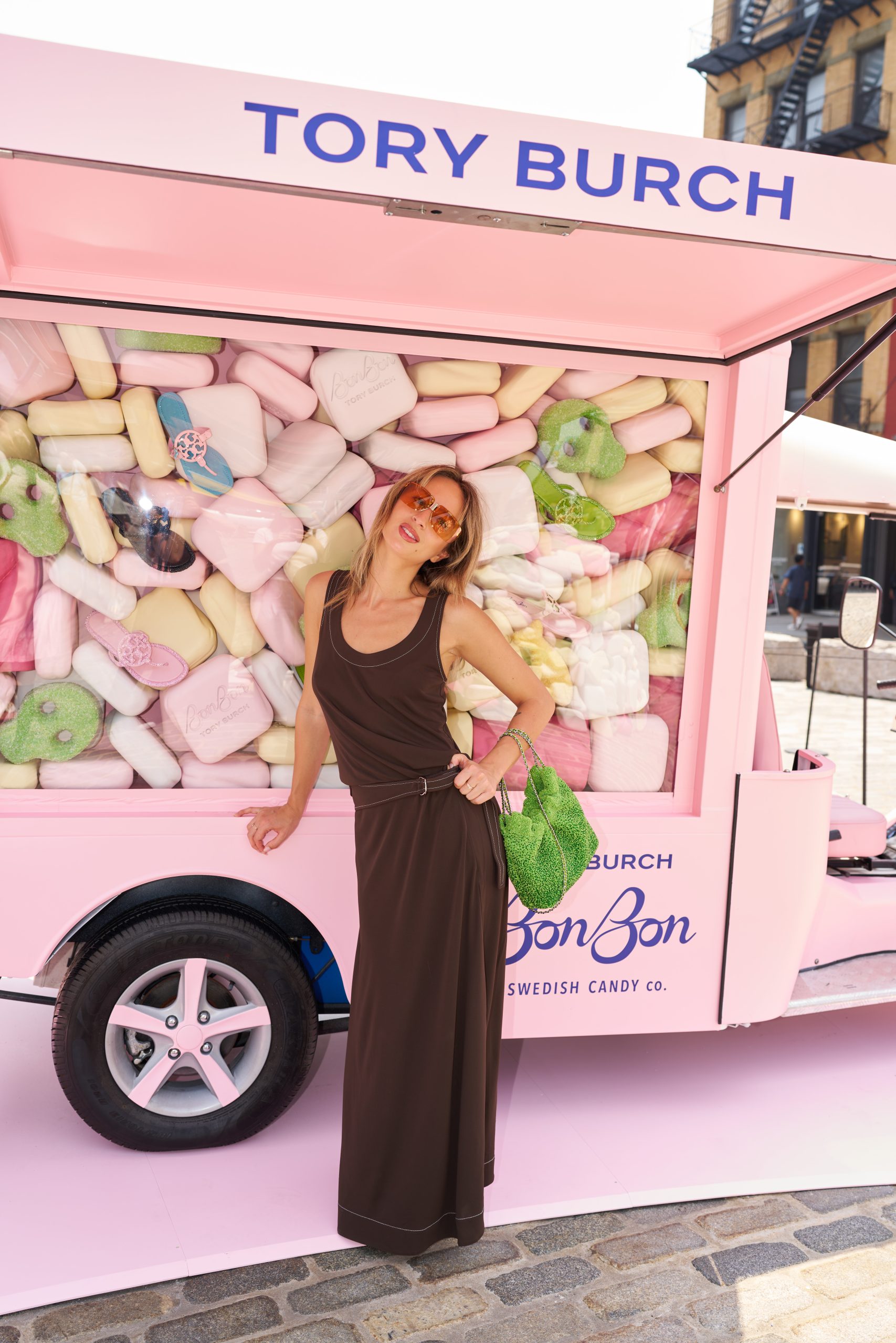 Woman in brown dress with red sunglasses and green handbag stands by pink candy truck with giant candies.