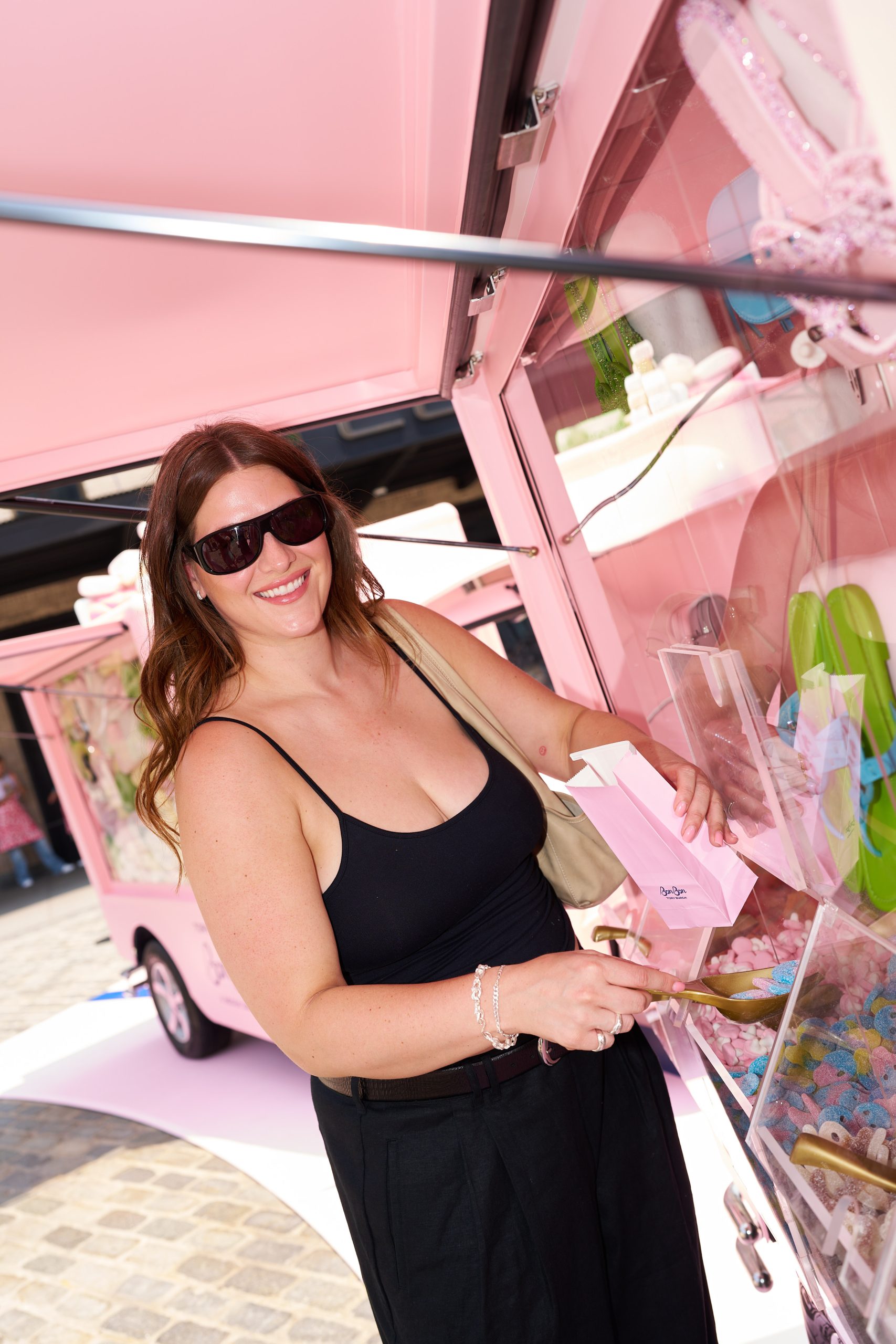 Woman in sunglasses and black tank top smiles while choosing candy from a pink food truck.