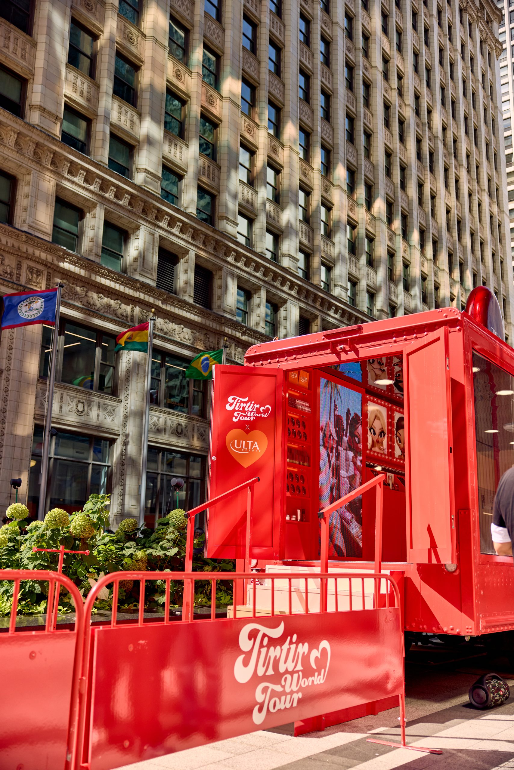 Red Tirtir World Tour pop-up booth on a city sidewalk, with office buildings and greenery nearby.