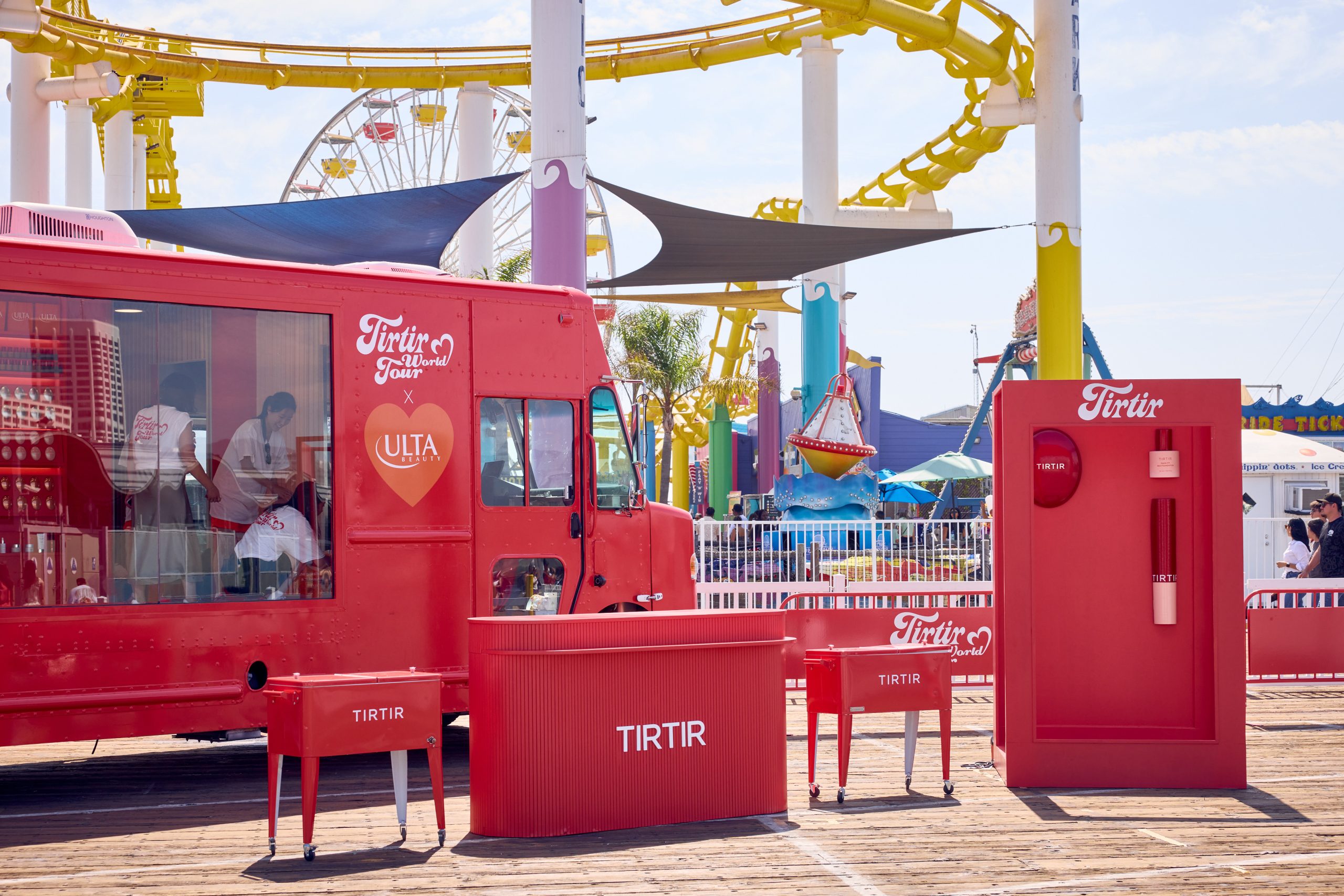 Red Tirtir and Ulta truck and booth outside near a roller coaster, ferris wheel, and people.