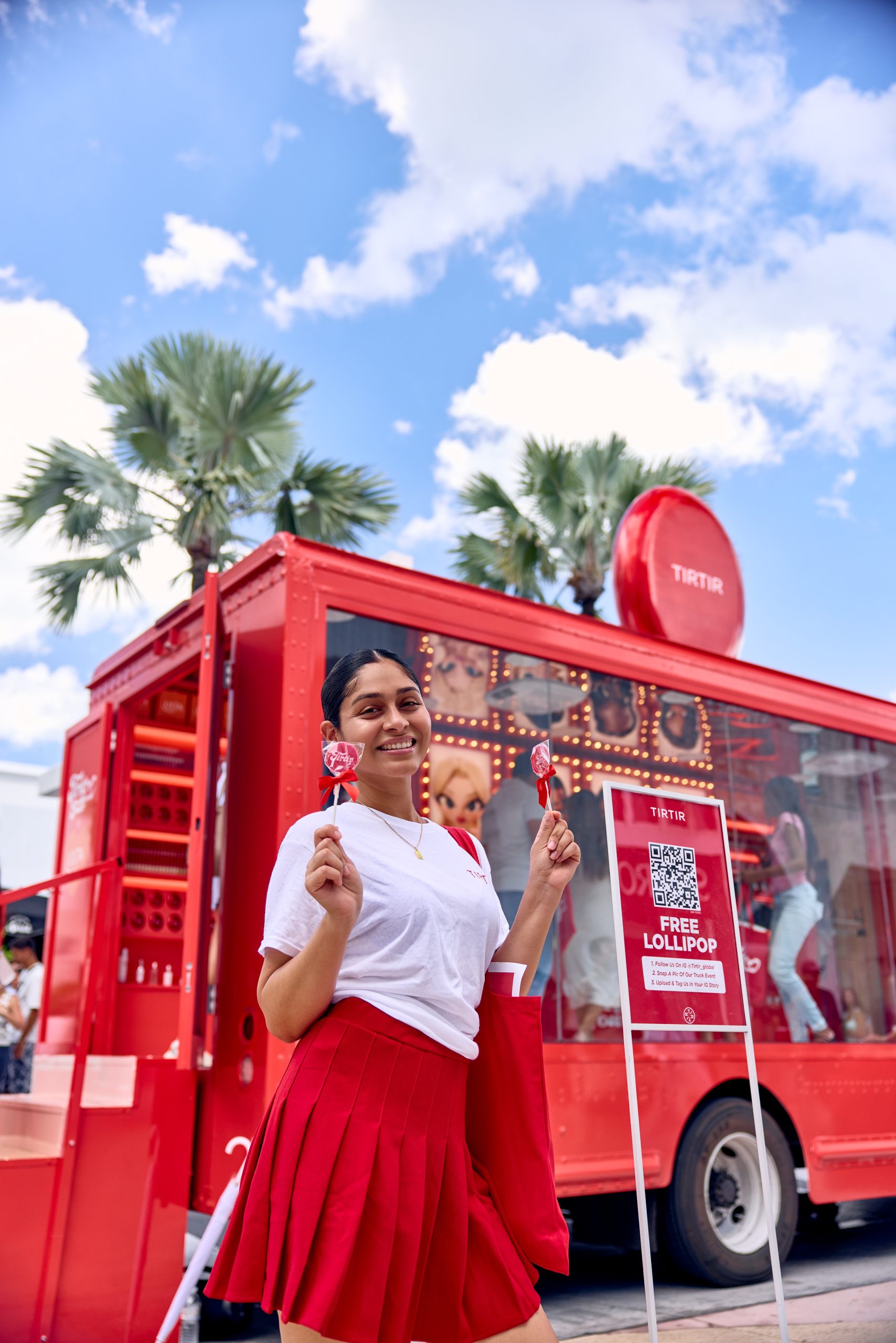 Woman in white shirt and red skirt holds two lollipops by a red promo truck with a sign for free lollipops.