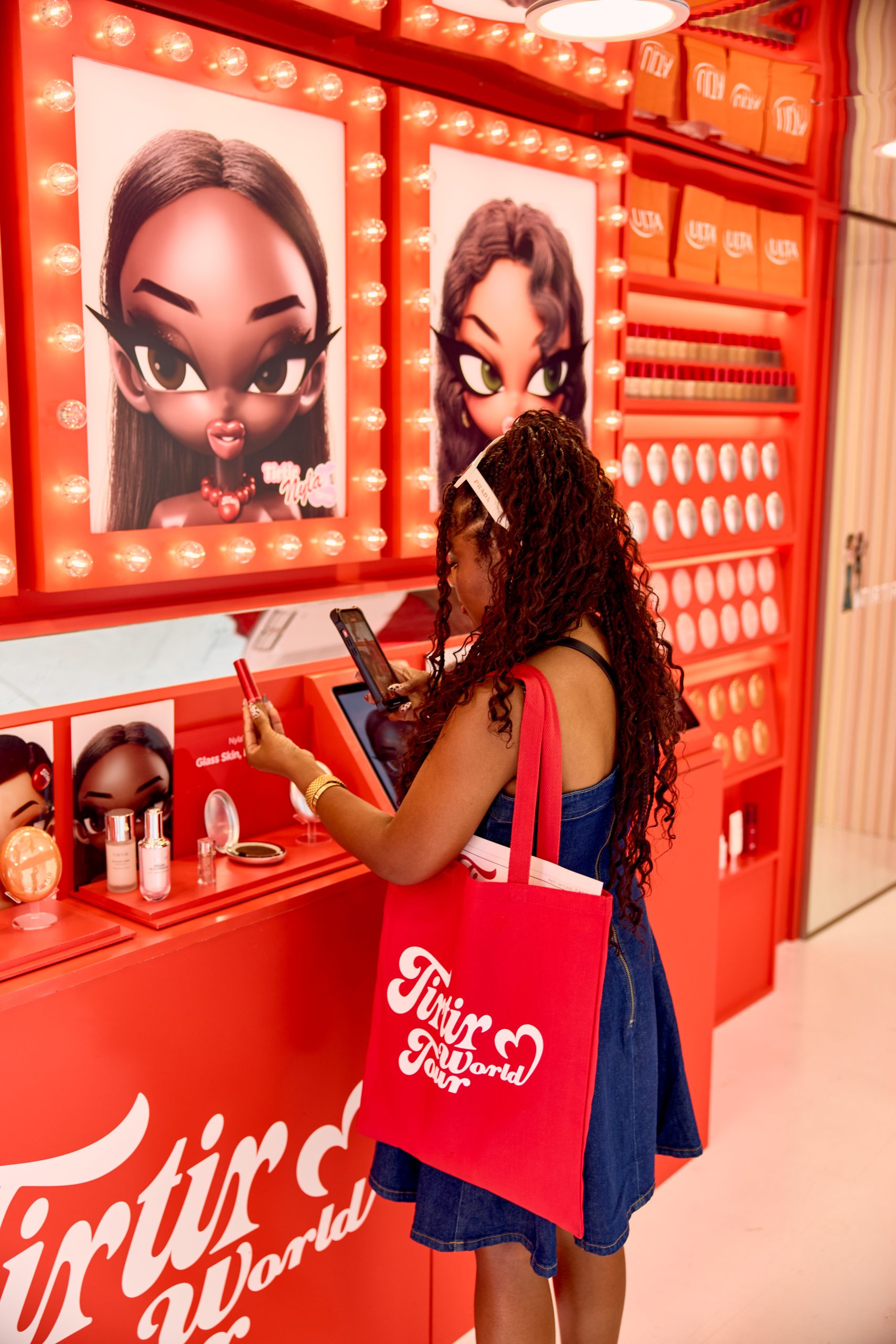 Woman with curly hair in blue dress and red tote bag looks at makeup at a display with red decor.