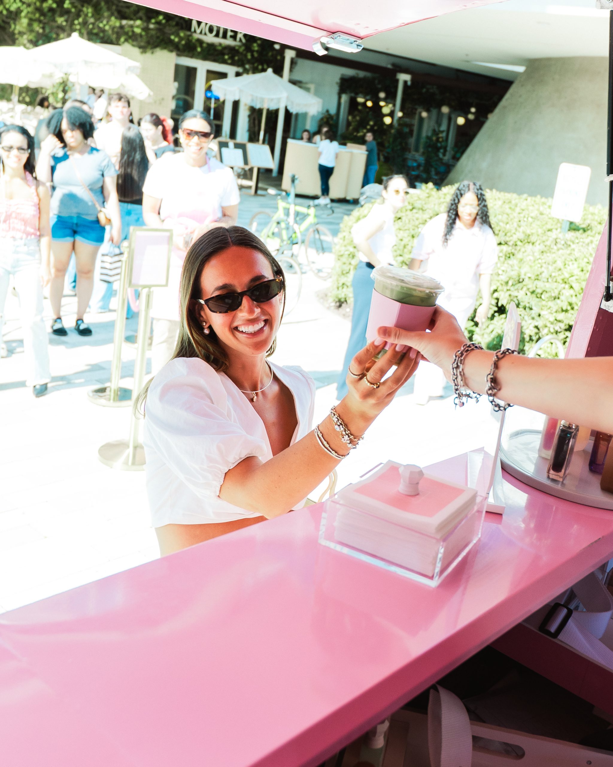 Woman in sunglasses gets a green drink from a pink food truck at an outdoor market.