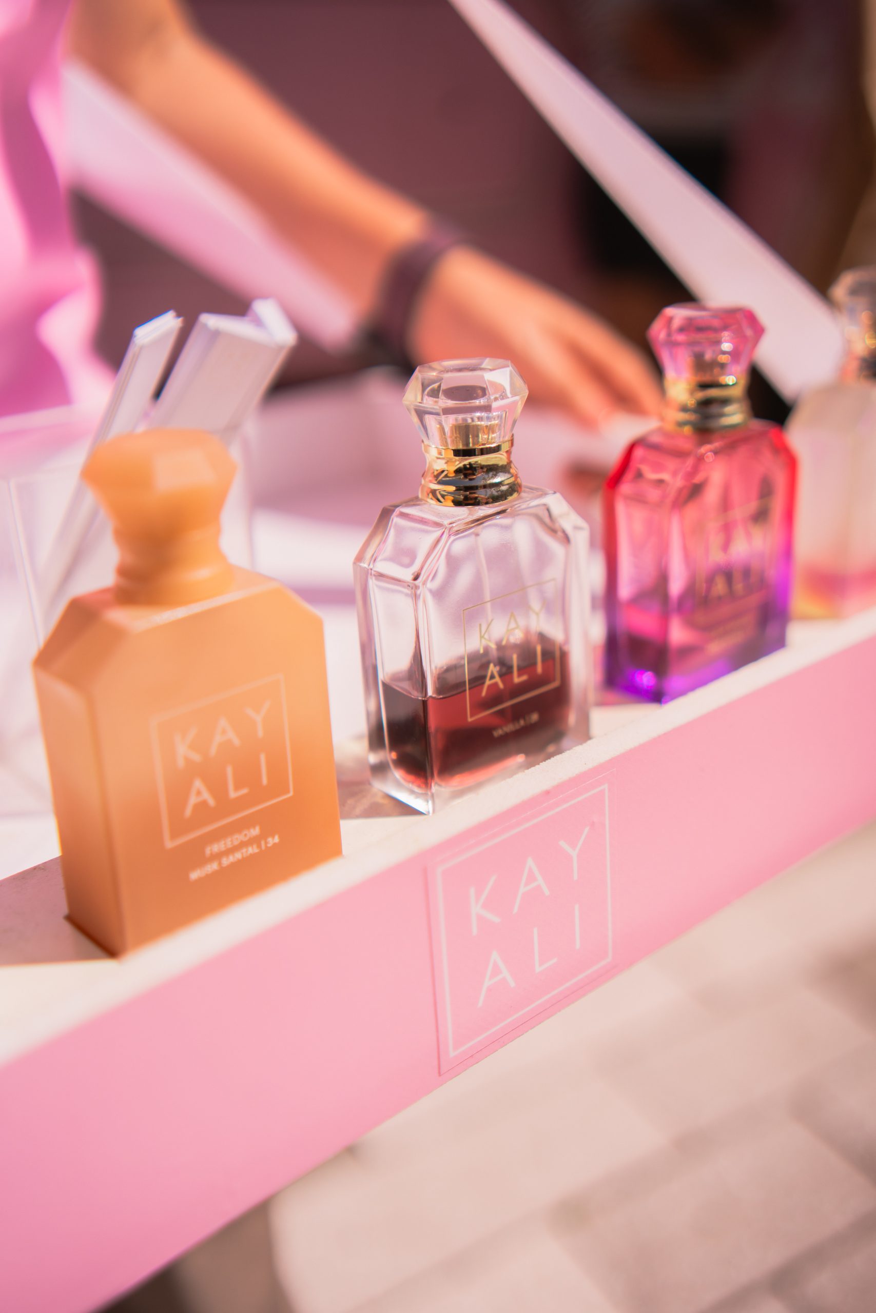 Four KAYALI perfume bottles in different colors sit on a pink stand, with an arm in the background.