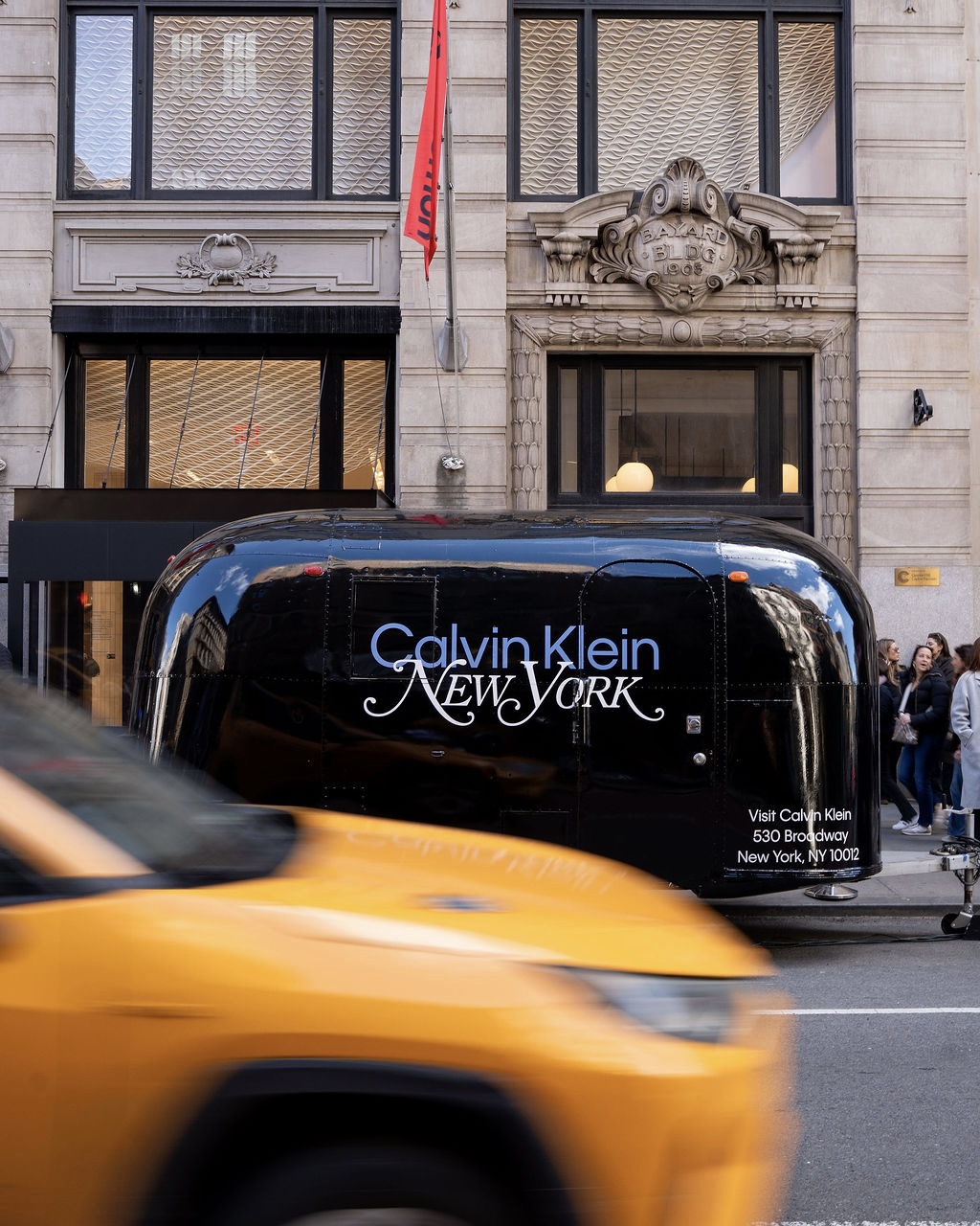 A yellow taxi drives by a Calvin Klein New York trailer outside a large building with people nearby.
