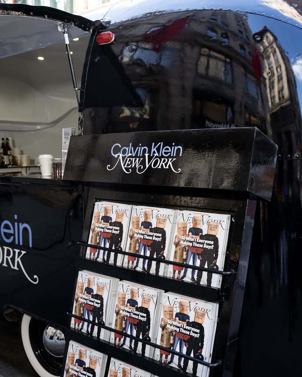 Magazine stand labeled Calvin Klein New York with Vogue magazines in front of a black trailer.
