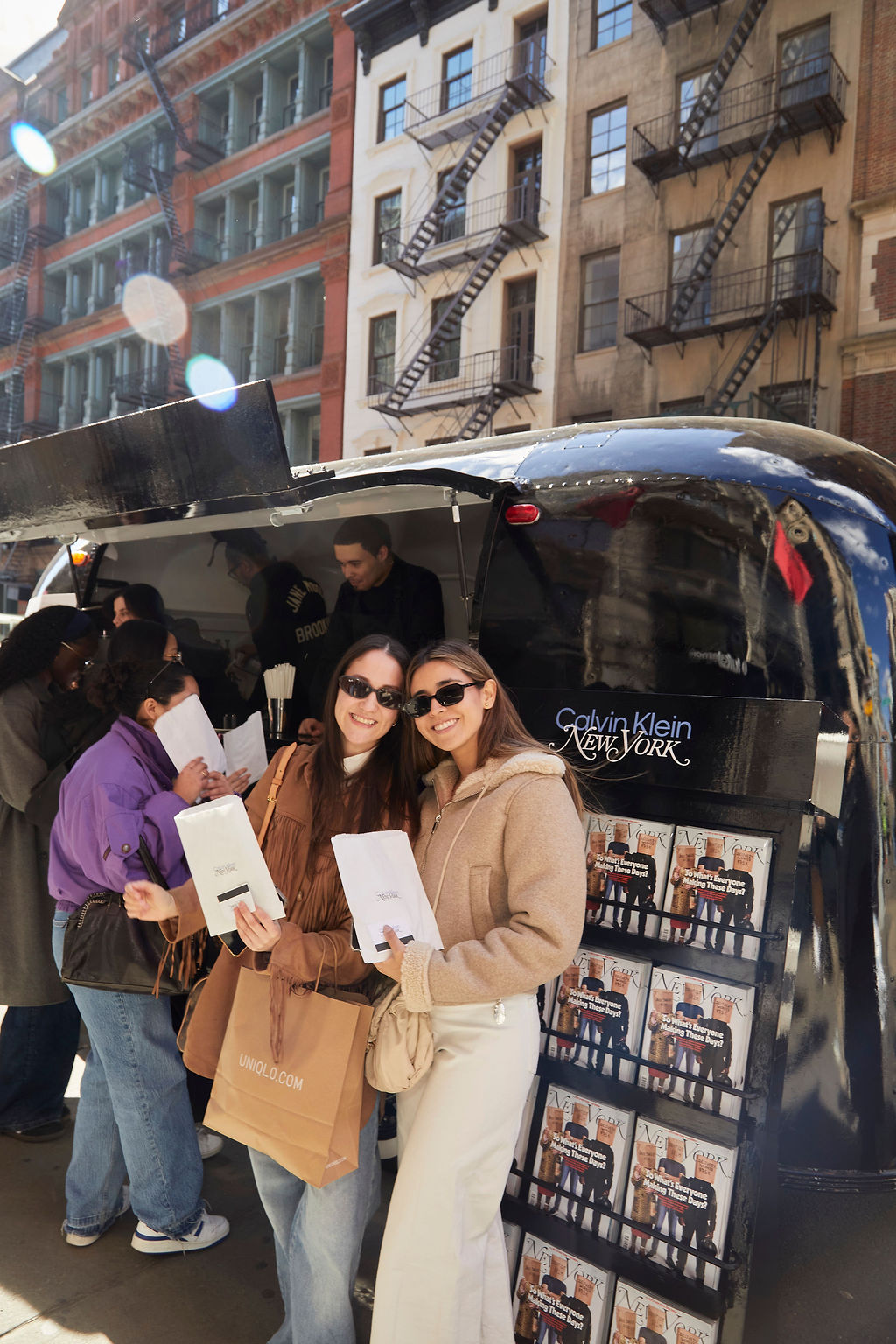 Two women in sunglasses hold paper bags by a Calvin Klein pop-up with people lining up behind them.