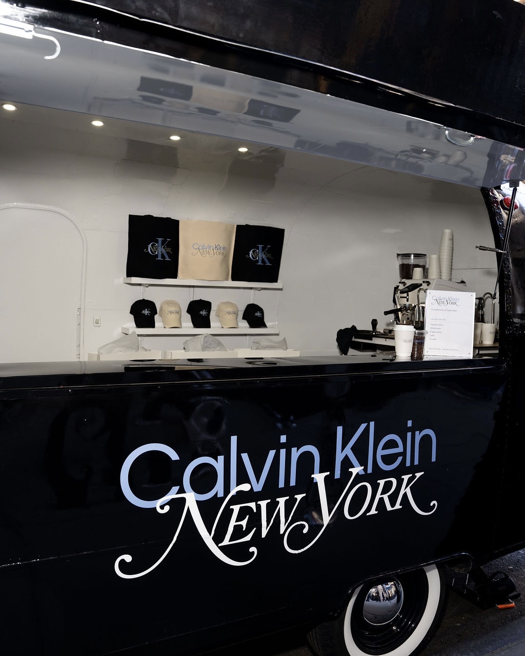 Black Calvin Klein food truck displaying branded hats, shirts, a coffee machine, and a menu.
