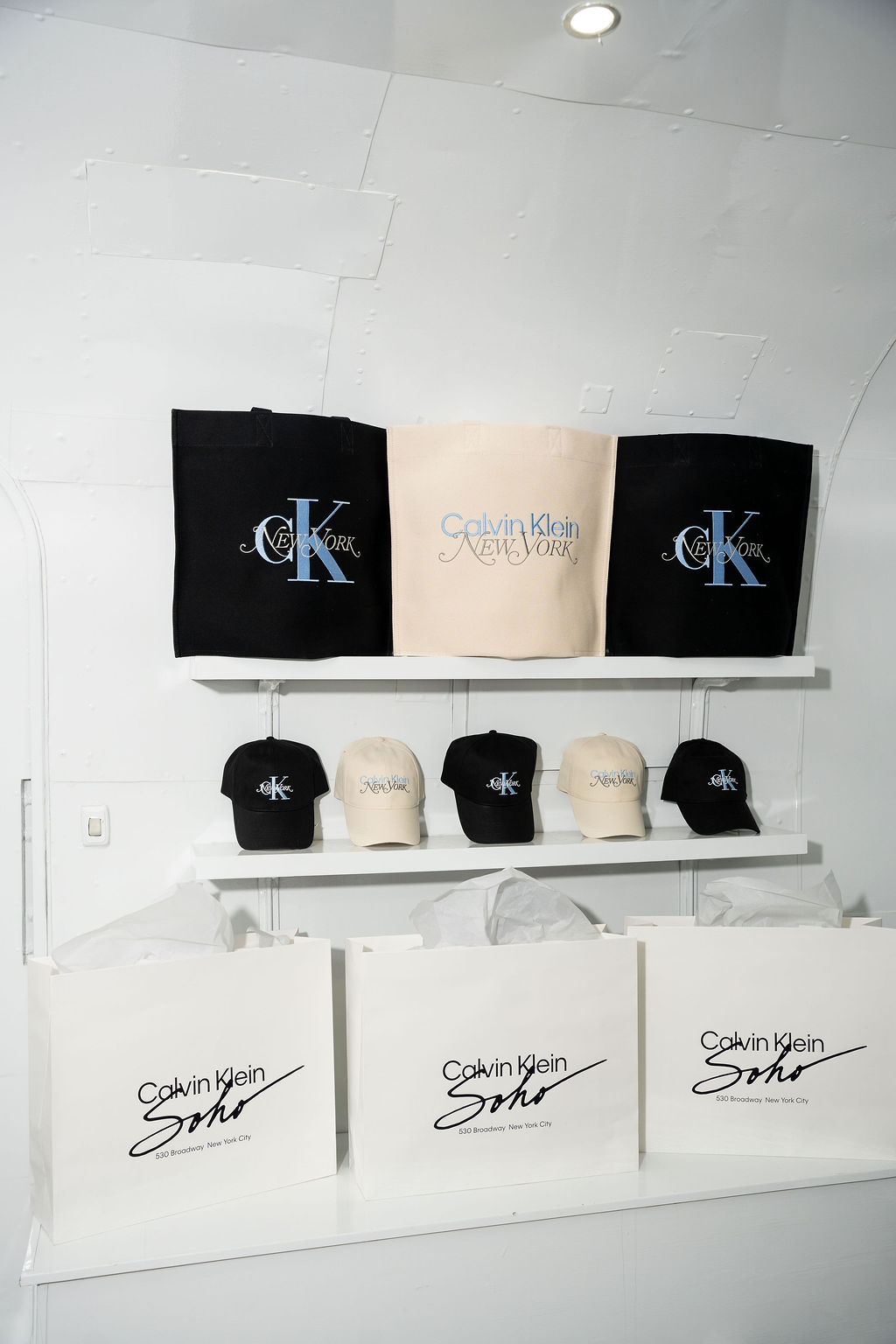 Calvin Klein tote bags and baseball caps in black and cream on shelves above shopping bags.