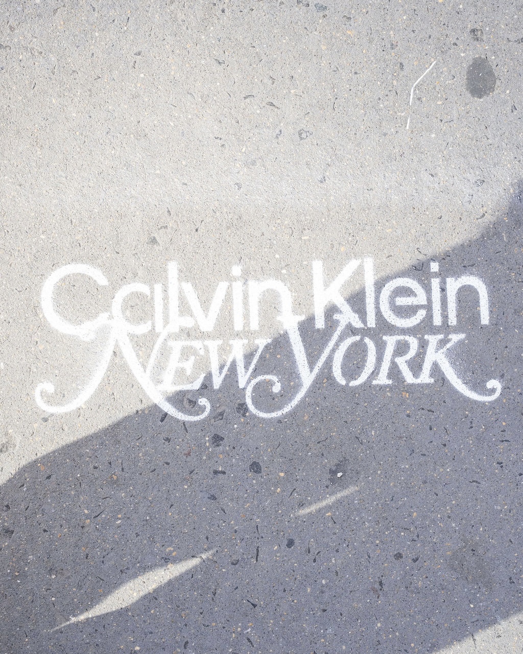 Calvin Klein NEW YORK is spray-painted in white on concrete, partly shaded and dusty.