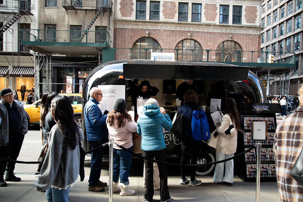 People line up at a black food truck on a city street with storefronts and a yellow taxi.