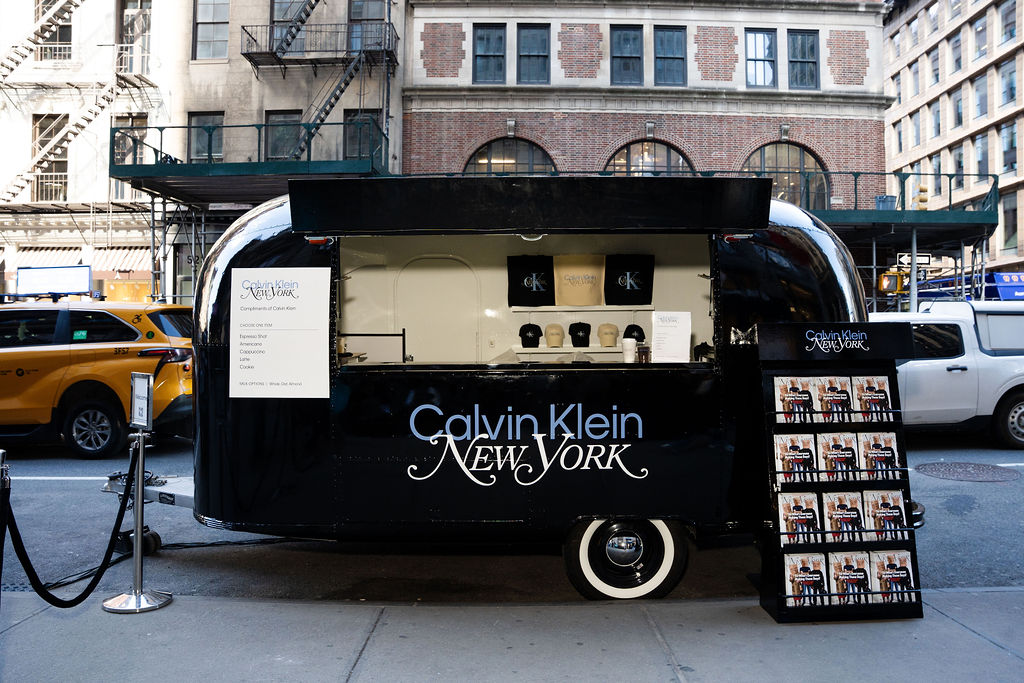 Black Calvin Klein trailer with open window displays hats; rack and yellow taxis in background.