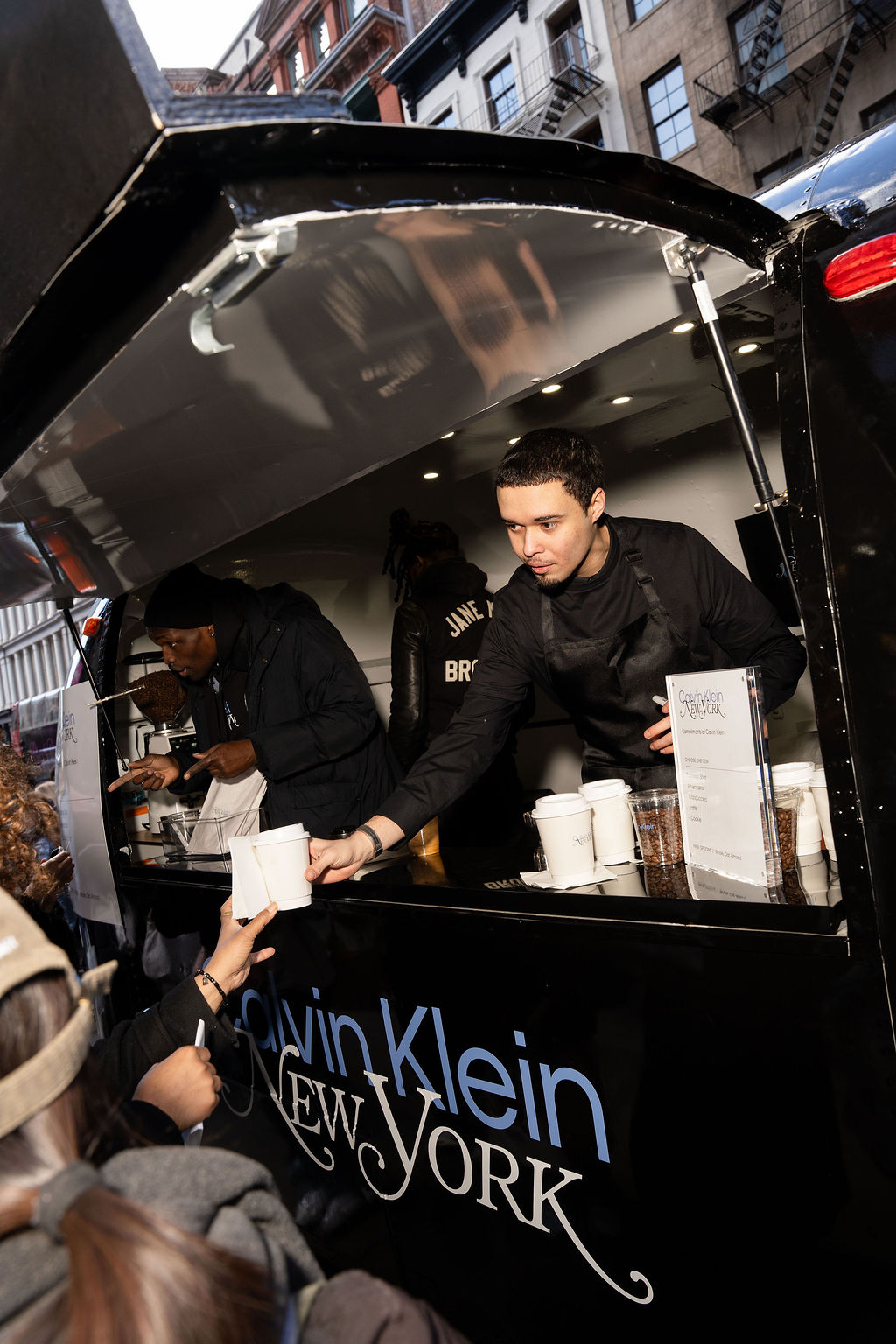 A Calvin Klein food truck worker hands a cup to a customer; the black truck shows the logo.