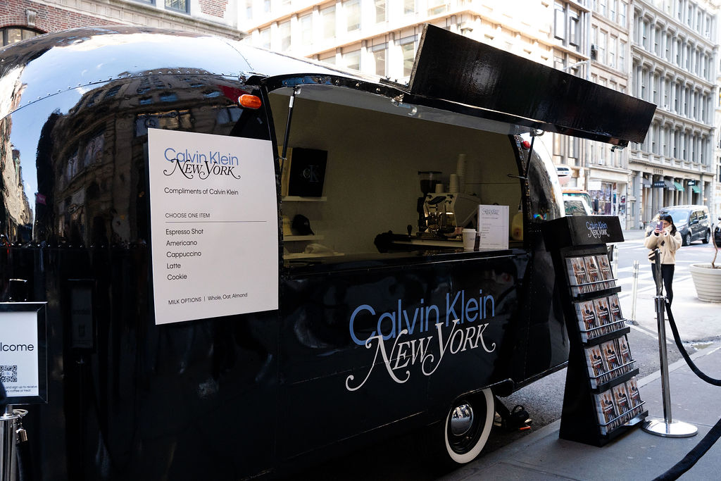 Calvin Klein coffee truck serves drinks on a city street with menu, brochures, people, and tall buildings.