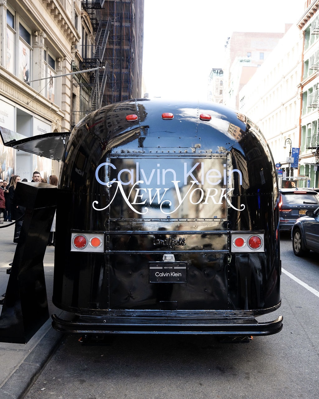 Black trailer with Calvin Klein NEW YORK on back parked on city street amid buildings and cars.