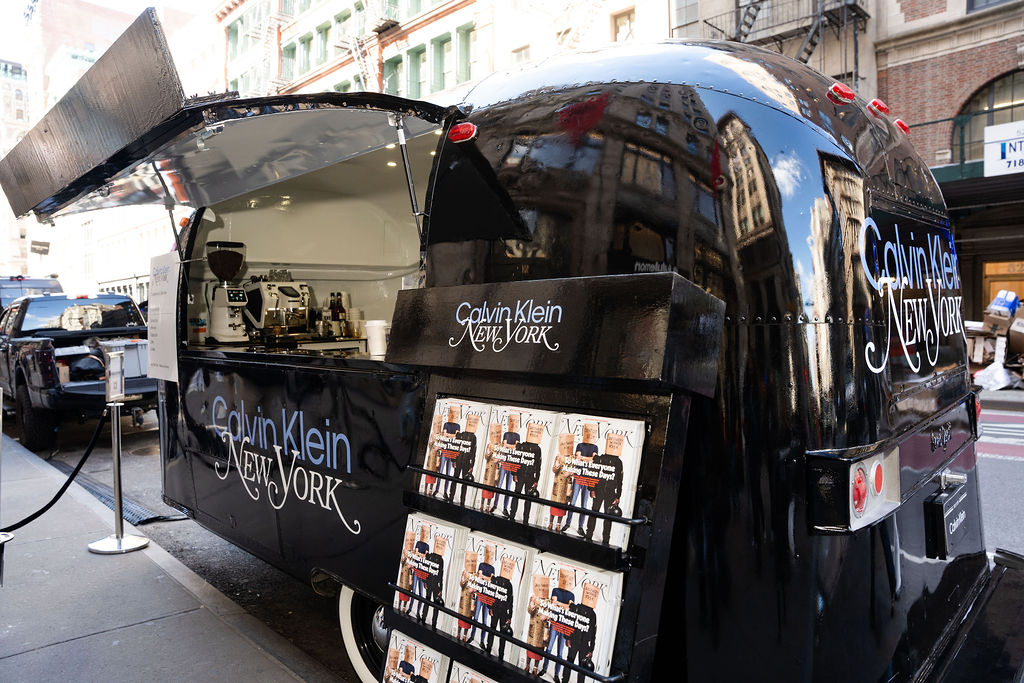 Calvin Klein branded food truck parked on city street with magazines displayed in front.