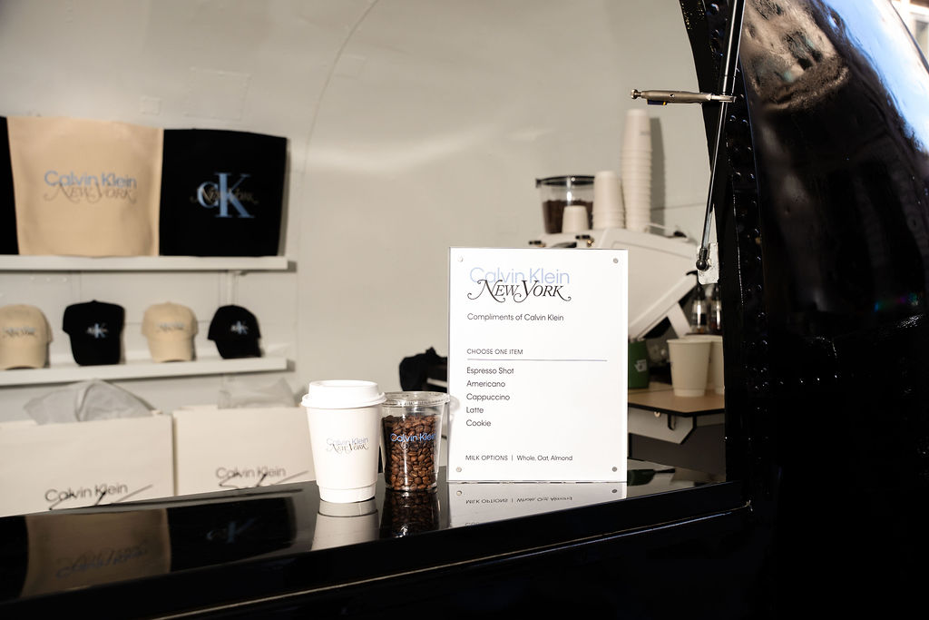 Coffee stand with Calvin Klein menu, cups, a jar of beans, and branded hats and bags in back.